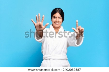 young hispanic woman smiling and looking friendly, showing number seven or seventh with hand forward, counting down. bathrobe concept