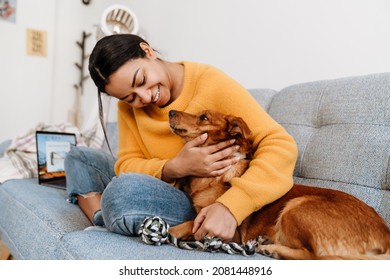 Young hispanic woman smiling and petting her dog while resting on couch at home