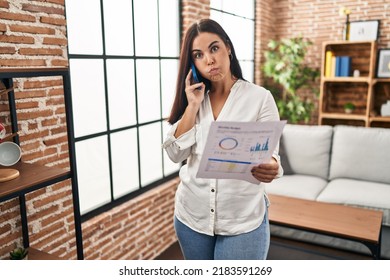 Young Hispanic Woman Speaking On The Phone About Bills Puffing Cheeks With Funny Face. Mouth Inflated With Air, Catching Air. 