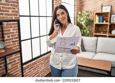 Young Hispanic Woman Speaking On The Phone About Bills Smiling Looking To The Side And Staring Away Thinking. 