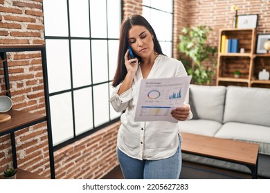 Young Hispanic Woman Speaking On The Phone About Bills Relaxed With Serious Expression On Face. Simple And Natural Looking At The Camera. 