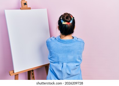 Young hispanic woman standing by painter easel stand standing backwards looking away and crossed arms 