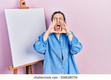 Young hispanic woman standing by painter easel stand shouting angry out loud and hands over mouth 