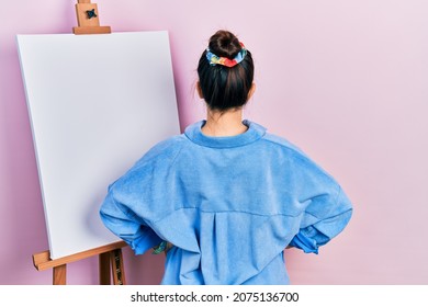 Young hispanic woman standing by painter easel stand standing backwards looking away and arms body 