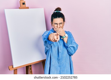 Young hispanic woman standing by painter easel stand laughing at you  pointing finger to the camera and hand over mouth  shame expression 