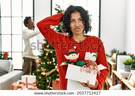 Young hispanic woman standing by christmas tree with decoration confuse and wondering about question. uncertain with doubt, thinking with hand on head. pensive concept. 