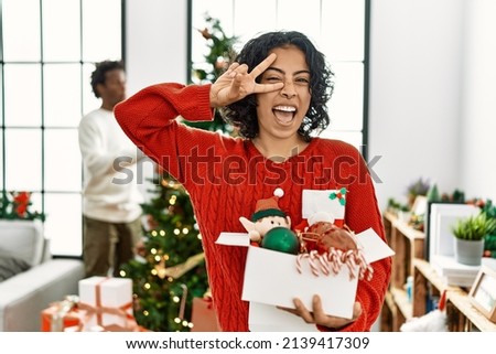 Young hispanic woman standing by christmas tree with decoration doing peace symbol with fingers over face, smiling cheerful showing victory 