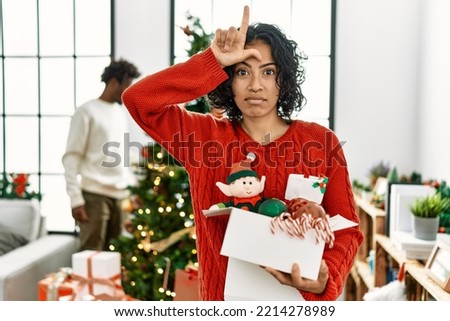 Young hispanic woman standing by christmas tree with decoration making fun of people with fingers on forehead doing loser gesture mocking and insulting. 