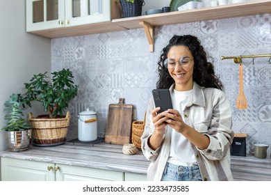 Young hispanic woman standing at home in kitchen with phone. Makes online purchases, orders, home delivery. - Powered by Shutterstock