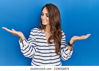 Young hispanic woman standing over blue isolated background smiling showing both hands open palms, presenting and advertising comparison and balance 