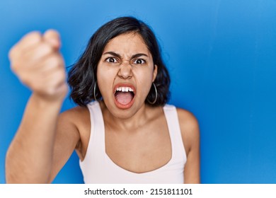 Young hispanic woman standing over blue background angry and mad raising fist frustrated and furious while shouting with anger. rage and aggressive concept. 