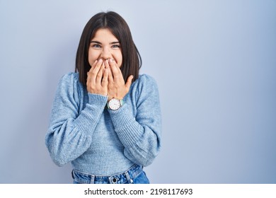 Young hispanic woman standing over blue background laughing and embarrassed giggle covering mouth with hands, gossip and scandal concept 