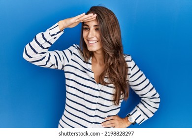Young hispanic woman standing over blue isolated background very happy and smiling looking far away with hand over head. searching concept. 
