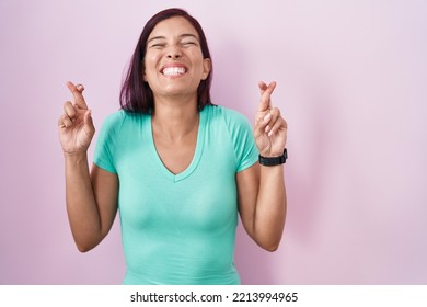 Young Hispanic Woman Standing Over Pink Background Gesturing Finger Crossed Smiling With Hope And Eyes Closed. Luck And Superstitious Concept. 