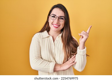 Young hispanic woman standing over yellow background with a big smile on face, pointing with hand and finger to the side looking at the camera.  - Shutterstock ID 2254769975