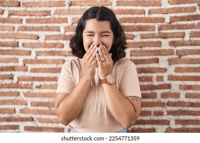 Young hispanic woman standing over bricks wall laughing and embarrassed giggle covering mouth with hands, gossip and scandal concept  - Shutterstock ID 2254771359