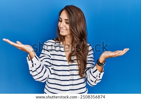 Young hispanic woman standing over blue isolated background smiling showing both hands open palms, presenting and advertising comparison and balance 