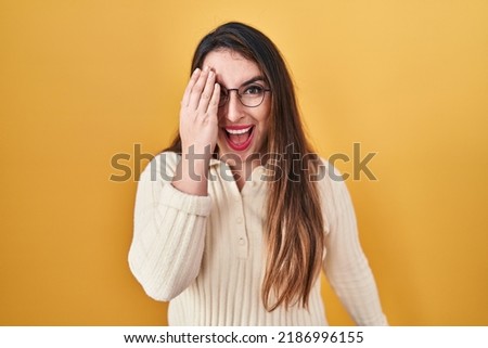Young hispanic woman standing over yellow background covering one eye with hand, confident smile on face and surprise emotion. 