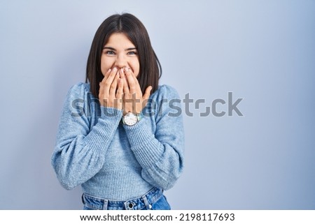 Young hispanic woman standing over blue background laughing and embarrassed giggle covering mouth with hands, gossip and scandal concept 