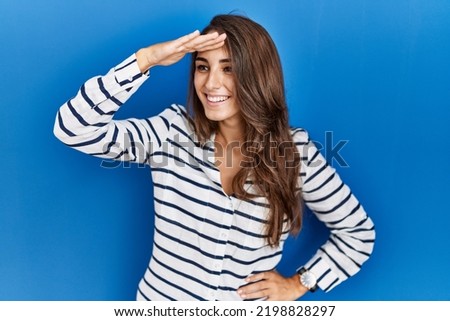 Young hispanic woman standing over blue isolated background very happy and smiling looking far away with hand over head. searching concept. 