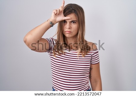 Young hispanic woman standing over isolated background making fun of people with fingers on forehead doing loser gesture mocking and insulting. 