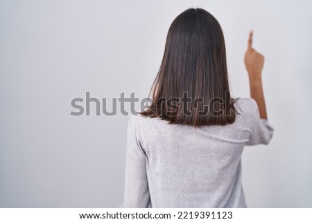 Young hispanic woman standing over white background posing backwards pointing ahead with finger hand 