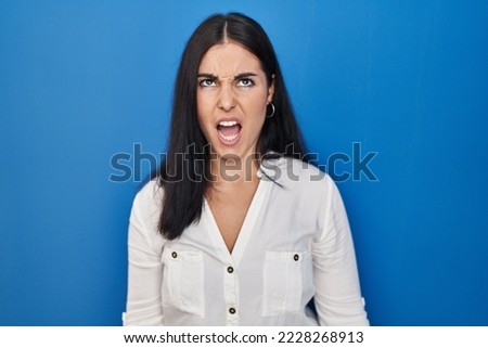 Young hispanic woman standing over blue background angry and mad screaming frustrated and furious, shouting with anger. rage and aggressive concept. 