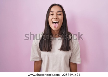 Young hispanic woman standing over pink background sticking tongue out happy with funny expression. emotion concept. 