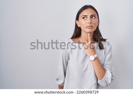 Young hispanic woman standing over white background thinking concentrated about doubt with finger on chin and looking up wondering 