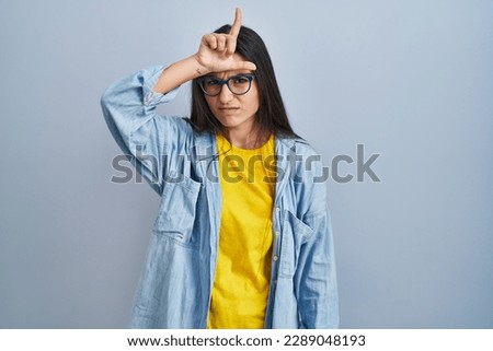 Young hispanic woman standing over blue background making fun of people with fingers on forehead doing loser gesture mocking and insulting. 