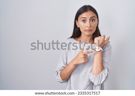 Young hispanic woman standing over white background in hurry pointing to watch time, impatience, looking at the camera with relaxed expression 