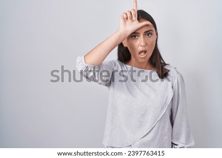 Young hispanic woman standing over white background making fun of people with fingers on forehead doing loser gesture mocking and insulting. 