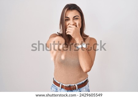 Young hispanic woman standing over white background laughing at you, pointing finger to the camera with hand over mouth, shame expression 