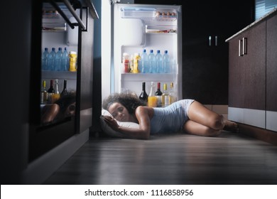 Junge hispanische Frau leidet an Sommerhitze und mangelnder Klimaanlage zu Hause. Schwarzes Mädchen mit Schweiß bedeckt schlafend auf dem Boden mit Kopf innen Kühlschrank.