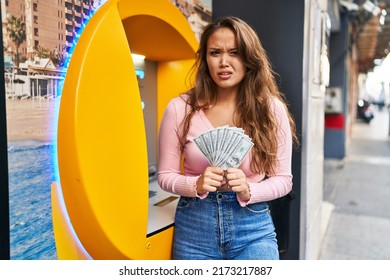 Young Hispanic Woman Taking Cash From Atm Clueless And Confused Expression. Doubt Concept. 