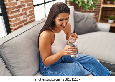 Young Hispanic Woman Taking Pill Drinking Water Sitting On Sofa At Home