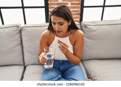 Young Hispanic Woman Taking Pill Drinking Water Sitting On Sofa At Home