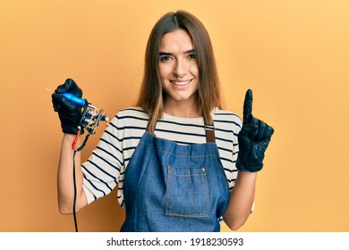 Young Hispanic Woman Tattoo Artist Wearing Professional Uniform And Gloves Smiling With An Idea Or Question Pointing Finger With Happy Face, Number One 