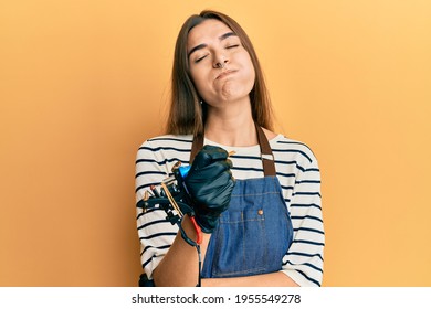 Young Hispanic Woman Tattoo Artist Wearing Professional Uniform And Gloves Puffing Cheeks With Funny Face. Mouth Inflated With Air, Catching Air. 