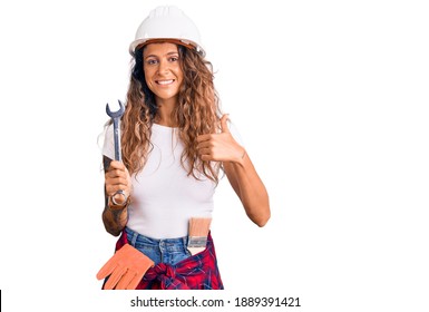 Young hispanic woman with tattoo wearing hardhat and builder clothes holding wrench smiling happy and positive, thumb up doing excellent and approval sign 
