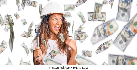 Young hispanic woman with tattoo wearing hardhat and builder clothes holding wrench pointing thumb up to the side smiling happy with open mouth