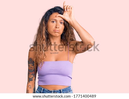 Young hispanic woman with tattoo wearing casual clothes making fun of people with fingers on forehead doing loser gesture mocking and insulting. 