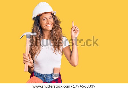 Young hispanic woman with tattoo wearing hardhat and builder clothes holding hammer surprised with an idea or question pointing finger with happy face, number one 