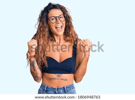 Young hispanic woman with tattoo wearing casual clothes and glasses celebrating surprised and amazed for success with arms raised and open eyes. winner concept. 