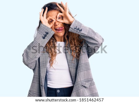 Young hispanic woman with tattoo wearing business oversize jacket doing ok gesture like binoculars sticking tongue out, eyes looking through fingers. crazy expression. 
