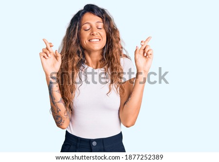 Young hispanic woman with tattoo wearing casual white tshirt gesturing finger crossed smiling with hope and eyes closed. luck and superstitious concept. 