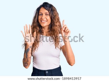 Young hispanic woman with tattoo wearing casual white tshirt showing and pointing up with fingers number six while smiling confident and happy. 