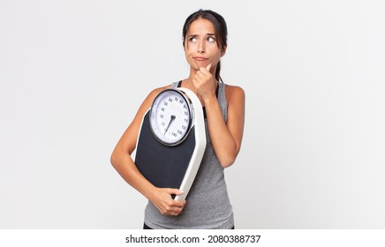 Young Hispanic Woman Thinking, Feeling Doubtful And Confused And Holding A Weight Scale. Diet Concept