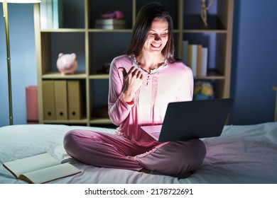 Young Hispanic Woman Using Computer Laptop On The Bed Pointing To The Back Behind With Hand And Thumbs Up, Smiling Confident 