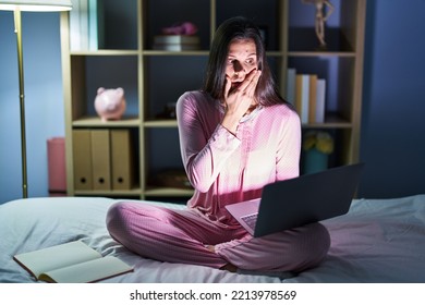 Young Hispanic Woman Using Computer Laptop On The Bed Shocked Covering Mouth With Hands For Mistake. Secret Concept. 